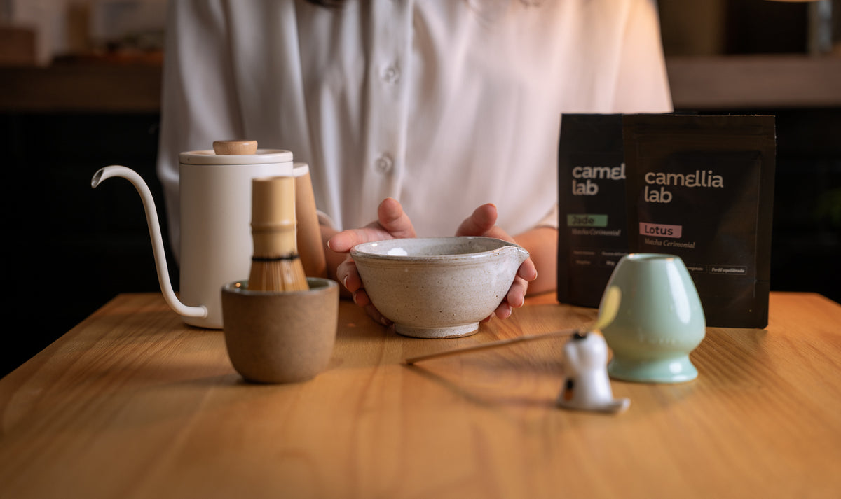 Mãos segurando uma tigela de cerâmica ao lado dos pacotes de matcha cerimonial Camellia Lab Lotus e Jade, com chaleira e batedor de bambu sobre mesa de madeira