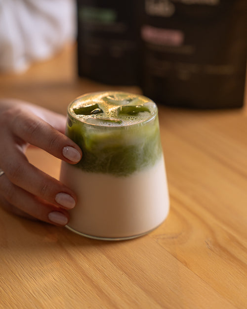Matcha cerimonial Lotus em latte gelado, com camada de matcha sobre leite e gelo, e os pacotes Camellia Lab ao fundo