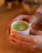 Matcha Lotus preparado quente, com espuma fina, em xícara de cerâmica segurada com as duas mãos